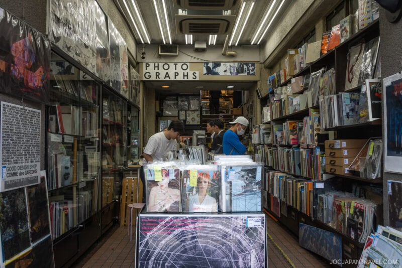 A small, narrow shop filled with books, magazines, and photo prints—a hidden gem among Tokyo bookstores. Several people browse the shelves beneath a PHOTOGRAPHS sign, while bright overhead lights illuminate this must-visit bookstore.