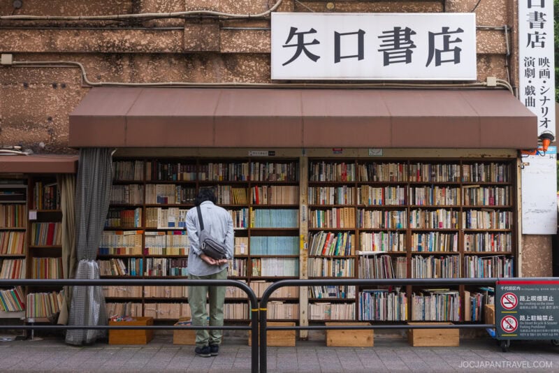 A person stands in front of an outdoor bookshop in Japan, browsing shelves filled with books. This charming spot is one of the must-visit bookstores in Tokyo, featuring a sign in Japanese and partially shaded by an awning.
