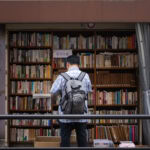 A man with a gray backpack stands in front of an outdoor bookshelf filled with books, browsing the selection—an inviting scene typical of must-visit bookstores in Tokyo, where shelves under brown awnings and boxes on the ground await discovery.