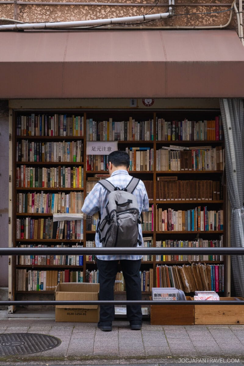 A man with a gray backpack stands in front of an outdoor bookshelf filled with books, browsing the selection&mdash;an inviting scene typical of must-visit bookstores in Tokyo, where shelves under brown awnings and boxes on the ground await discovery.