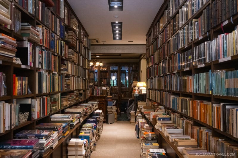 A cozy, narrow bookstore with tall shelves packed with books on both sides and stacks on the floor. Warm lighting and a lamp create an inviting atmosphere. A table and chairs in the back make it one of the must-visit bookstores in Tokyo.