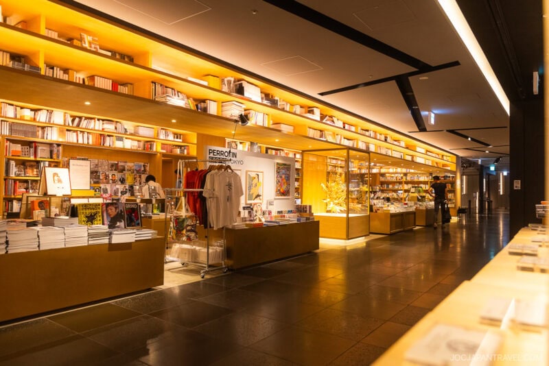 A brightly lit bookstore with wooden shelves filled with books, magazines, and merchandise, including t-shirts. Among must-visit bookstores, its modern design and warm atmosphere make it a standout choice for anyone exploring Tokyo bookstores.
