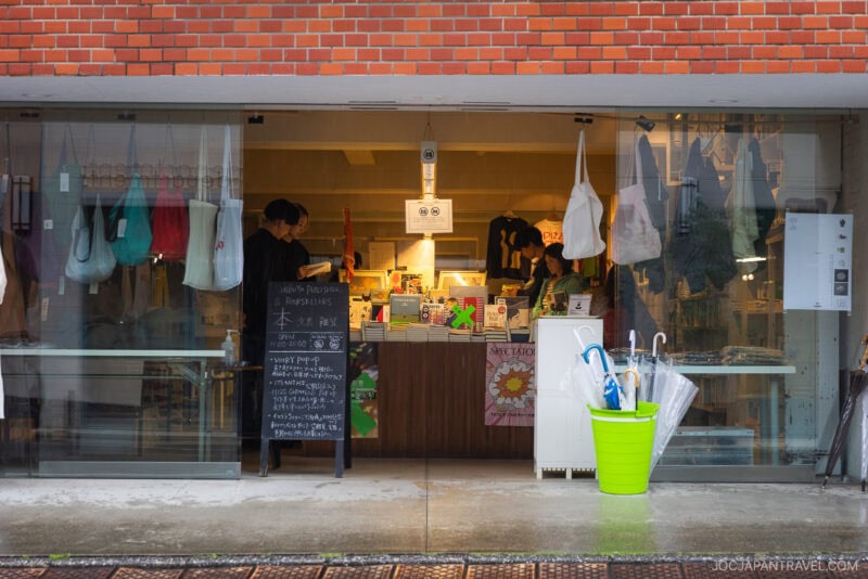 A small shop with large glass windows displays tote bags, books, and various items—a scene typical of must-visit bookstores in Tokyo. Two people stand behind the counter, while a green bucket holding umbrellas sits outside in the rain.