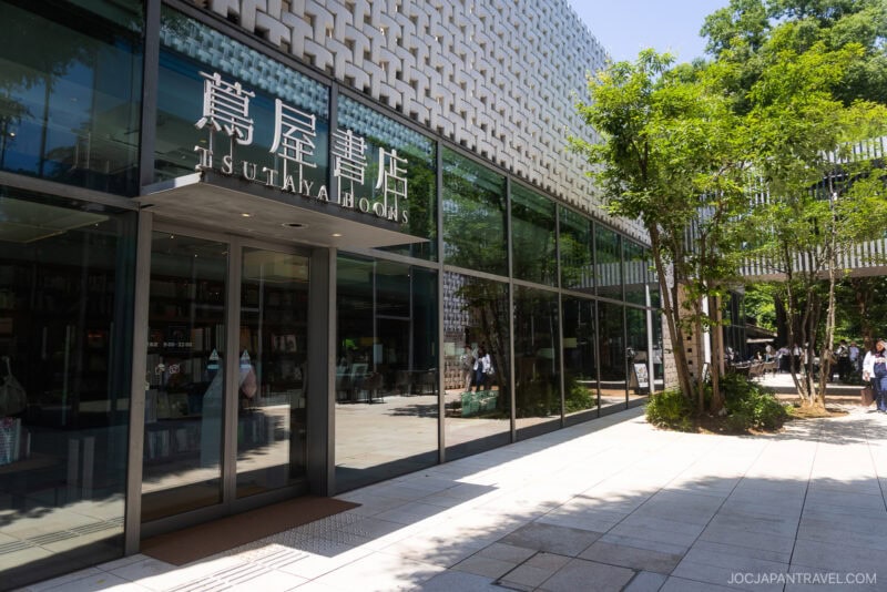 The glass exterior of Tsutaya Books in Daikanyama, one of the must-visit bookstores in Tokyo, is surrounded by trees along a sunlit walkway, with shelves of books visible through the large windows.