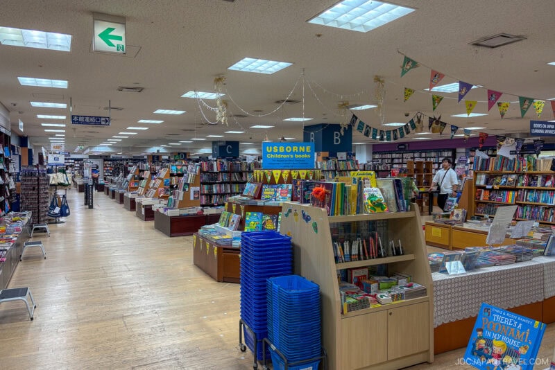 A spacious bookstore interior in Tokyo with shelves of books, colorful childrens books on display, blue baskets stacked in the aisle, and a sign reading Usborne Childrens Books—a must-visit bookstore for any book lover. A person browses in the background.