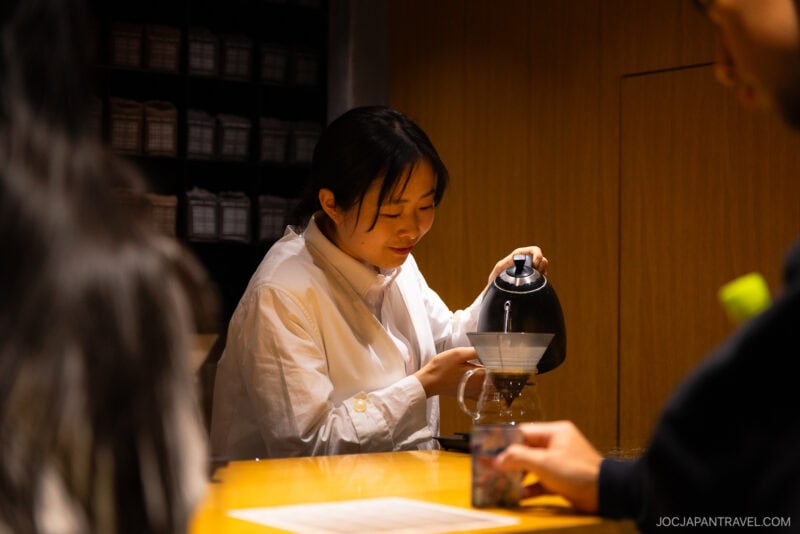 A woman in a white shirt is carefully pouring hot water from a kettle into a pour-over coffee dripper at a wooden counter, capturing the meticulous craft found in must-visit cafes and Tokyo coffee shops. Shelves and other people are seen in the background.