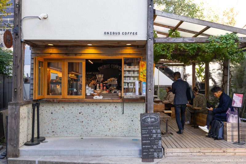Street view of Onibus Coffee, one of the best cafes Tokyo offers, featuring a takeout window, outdoor seating, people relaxing and ordering, and a menu board by the entrance under a pergola with greenery—perfect for any Tokyo cafe guide.