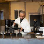 A barista in a white coat pours hot water into coffee drippers behind a modern café counter, capturing the artisanal spirit often found in the best cafes Tokyo has to offer. Coffee equipment and boxes are visible in the background.