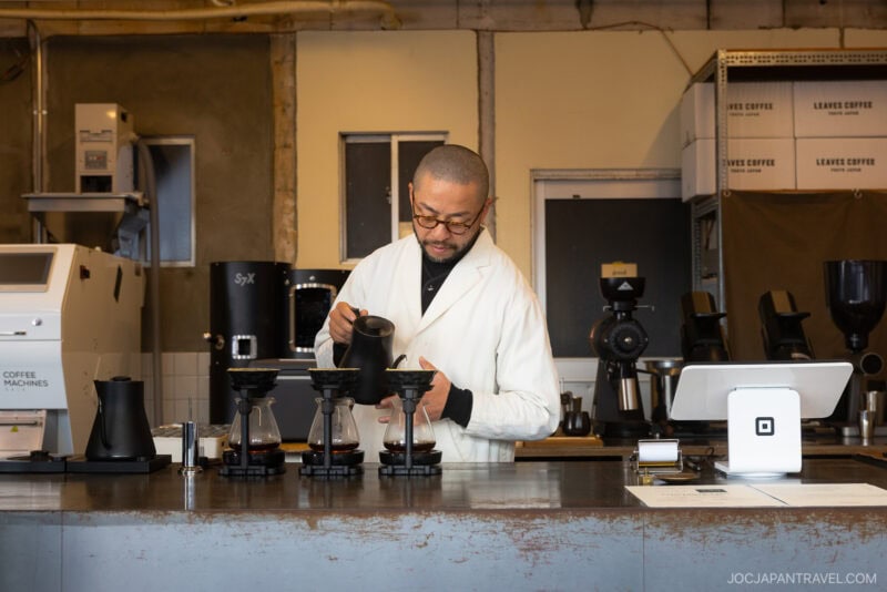 A barista in a white coat pours hot water into coffee drippers behind a modern caf&eacute; counter, capturing the artisanal spirit often found in the best cafes Tokyo has to offer. Coffee equipment and boxes are visible in the background.