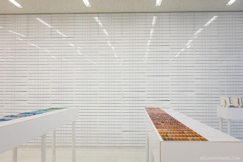 A minimalist white room with wall-to-wall drawers labeled in Japanese; display tables in the foreground showcase organized colored pencils, pens, and art supplies—a scene reminiscent of stationery stores Tokyo is known for.