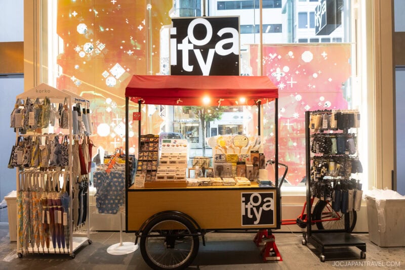 A small wooden cart with a red canopy displays Japanese paper, postcards, and fans in a brightly lit indoor space. On either side are racks with patterned umbrellas and pens. A “toaty” sign evokes the charm of stationery stores Tokyo is known for.