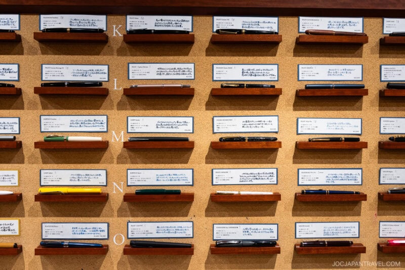 A display wall featuring rows of Japanese pens on wooden shelves, each with a handwritten label above it. Letters K, L, M, and N mark the rows on the left side of the corkboard background—true to stationery stores Tokyo style.
