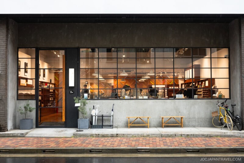 A modern bookstore with large glass windows, wooden shelves filled with books, a bench, potted plants, and a yellow bicycle parked outside on a wet sidewalk—reminiscent of stationery stores Tokyo, known for Japanese pens and fine Japanese paper.