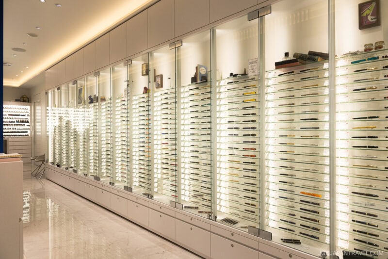 A well-lit, modern display case with multiple shelves showcases an extensive collection of Japanese pens in a sleek, white showroom with glossy marble floors.
