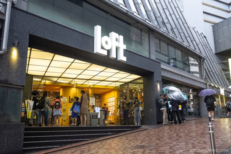 People with umbrellas stand outside and enter a brightly lit Loft store—one of the top stationery stores Tokyo offers—on a rainy day in an urban area, with the store’s large white sign visible above the entrance.