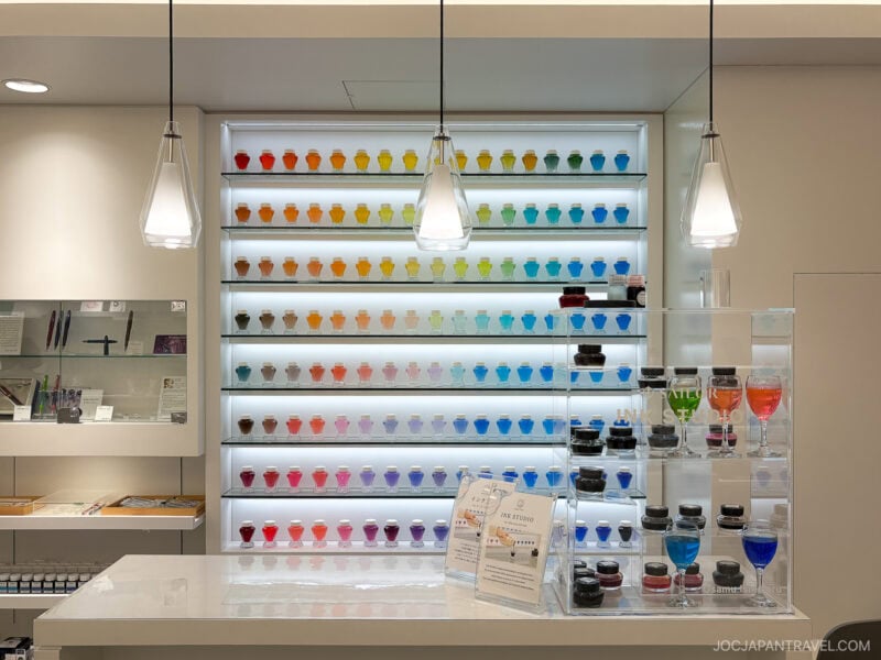 A brightly lit store display features shelves of colorful ink bottles arranged in a rainbow gradient behind a white counter, with Japanese pens and sample inks on the counter&mdash;reminiscent of the finest stationery stores Tokyo has to offer.