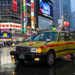 Things to do on a Rainy Day in Tokyo A yellow taxi with its door open is parked on a rainy street in a busy city area at night, surrounded by neon signs, tall buildings, and crowds—capturing the vibrant energy of Tokyo activities, even on a rainy day.