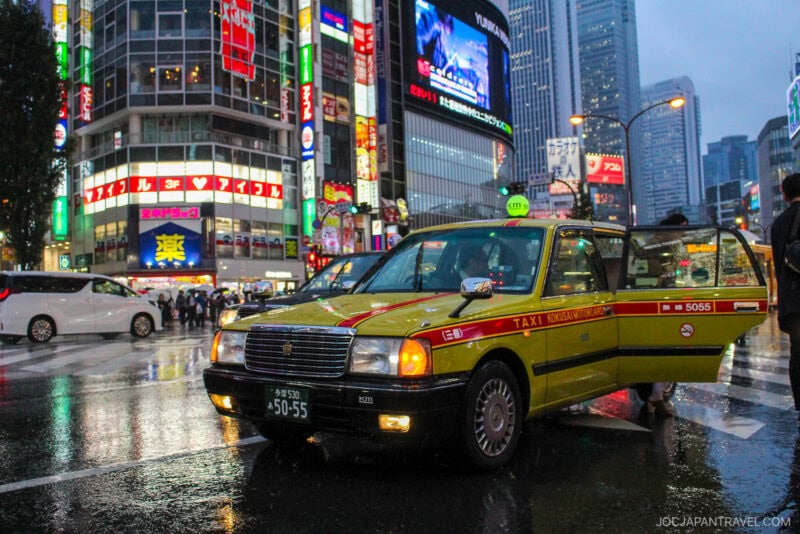 A yellow taxi with its door open is parked on a rainy street in a busy city area at night, surrounded by neon signs, tall buildings, and crowds&mdash;capturing the vibrant energy of Tokyo activities, even on a rainy day.