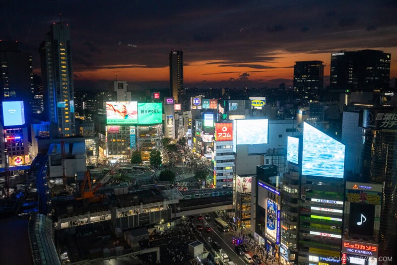 A vibrant cityscape of Shibuya, Tokyo at dusk, with illuminated billboards and busy streets—perfect for discovering things to do Tokyo even as rain begins to fall under a dramatic orange and blue sunset sky.