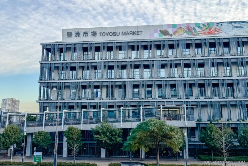 A modern multi-story building with large windows and a sign that reads “Toyosu Market” in English and Japanese, featuring seafood illustrations above. Trees line the sidewalk outside Tokyo’s famous fish market, under a partly cloudy sky.