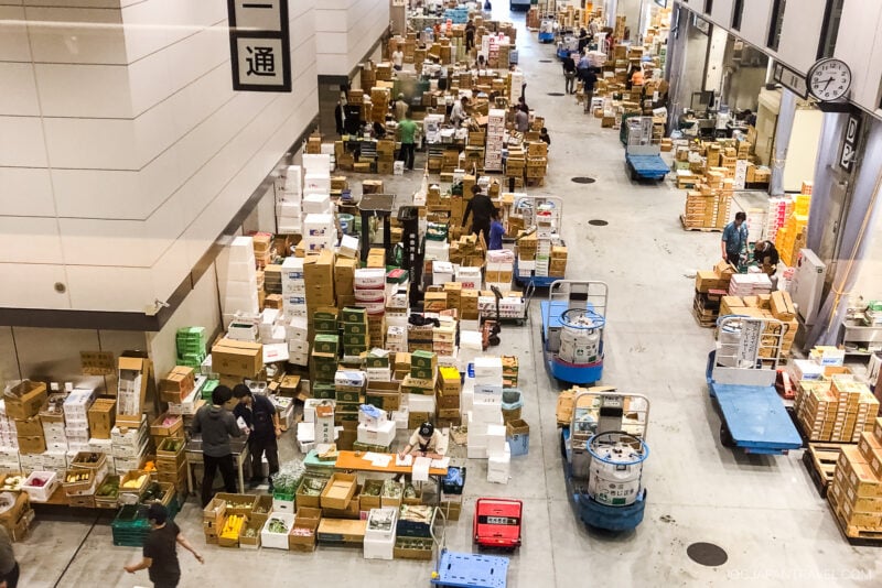 A bustling indoor market reminiscent of Toyosu Market, with people sorting and organizing stacks of boxes and crates, carts, and various goods—capturing the lively energy of a Tokyo fish market during a seafood auction.