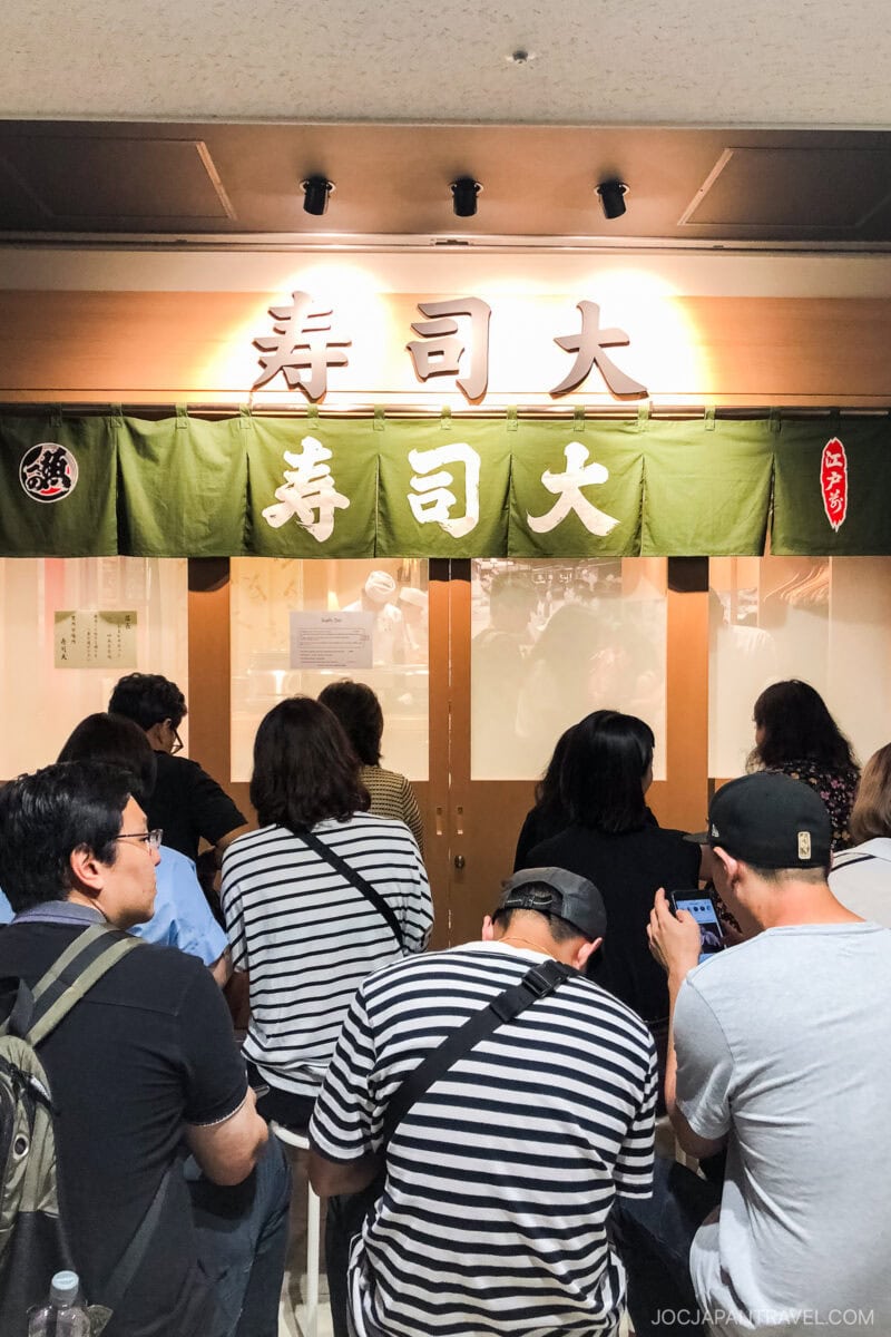 A group of people sit and wait in front of a Japanese sushi restaurant with a green curtain and white kanji characters above the entrance, reminiscent of eateries near Toyosu Market, famed for its Tokyo Fish Market seafood auction.