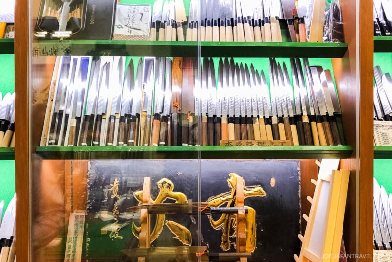 A display case filled with various Japanese kitchen knives, neatly arranged in rows on glass shelves, with a decorative plaque featuring gold Japanese characters below—much like those seen at the renowned Toyosu Market in Tokyo’s famous fish market.