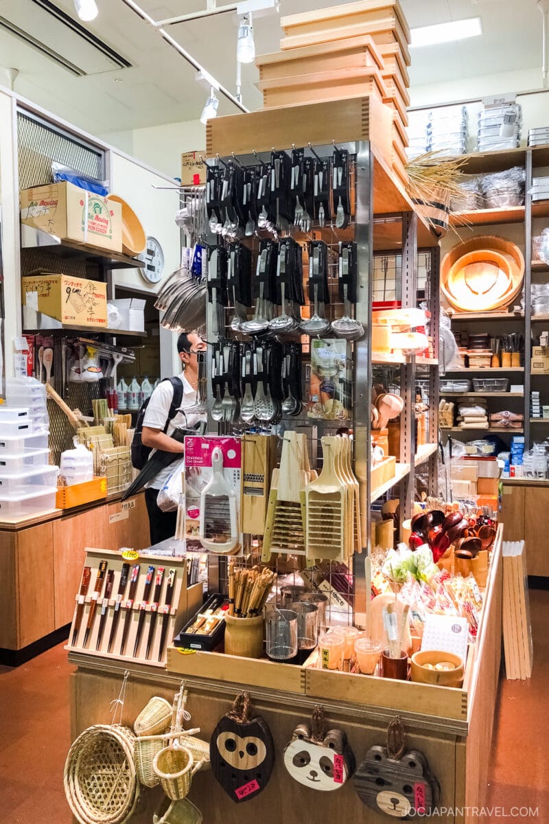 A store display filled with various kitchen utensils, baskets, brushes, and wooden tools evokes the vibrant energy of the Tokyo fish market. A masked shopper browses shelves under bright lights and neatly organized items.