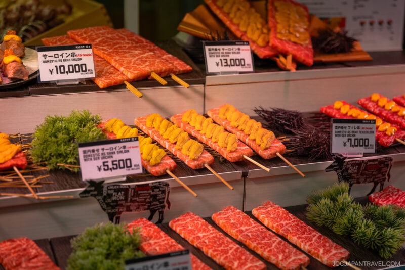 Display case of premium marbled beef cuts and skewers topped with sea urchin at Tsukiji Outer Market, with price tags in Japanese yen and decorative garnishes.