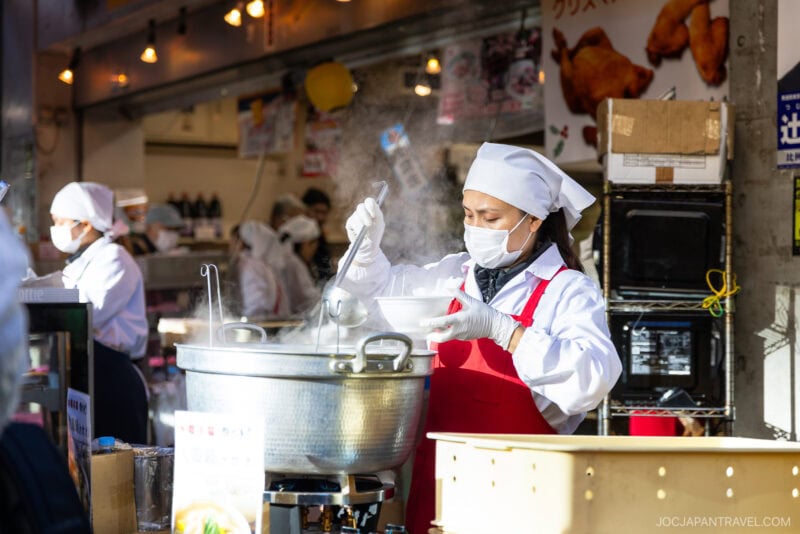 At Tokyo’s busy Tsukiji Outer Market, a woman in a white chef’s hat, mask, and red apron ladles steaming soup into a bowl at her bustling food stall, with other workers moving in the background.