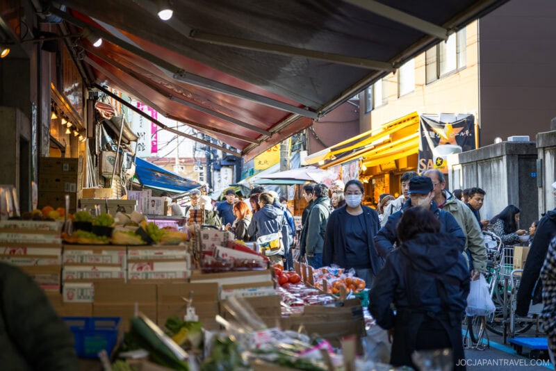 A busy outdoor market in Tokyo with shoppers walking among fruit and vegetable stalls at Tsukiji Outer Market, some wearing masks. Colorful produce fills boxes, while various shop signs add to the lively scene in the background.