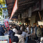 Tokyo Tsukiji Outer Market A busy outdoor market scene at Tokyo’s Tsukiji Outer Market, with many people walking and browsing stalls under an awning; shops and signs with Japanese writing line the street as sunlight filters through trees on the left.
