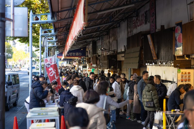 A busy outdoor market scene at Tokyo&rsquo;s Tsukiji Outer Market, with many people walking and browsing stalls under an awning; shops and signs with Japanese writing line the street as sunlight filters through trees on the left.