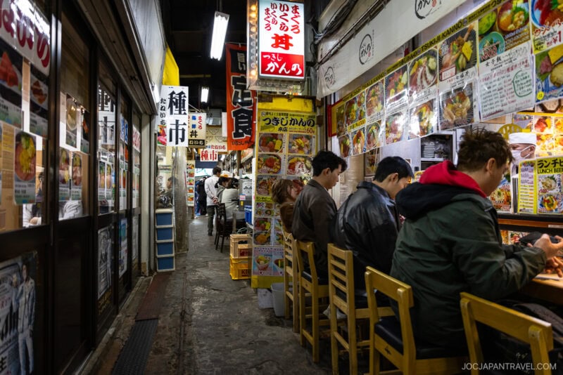 A narrow alleyway in Tokyo’s Tsukiji Outer Market with people sitting on stools eating at a small restaurant, surrounded by colorful food menus and signs with Japanese text. Bright lights illuminate the bustling scene.