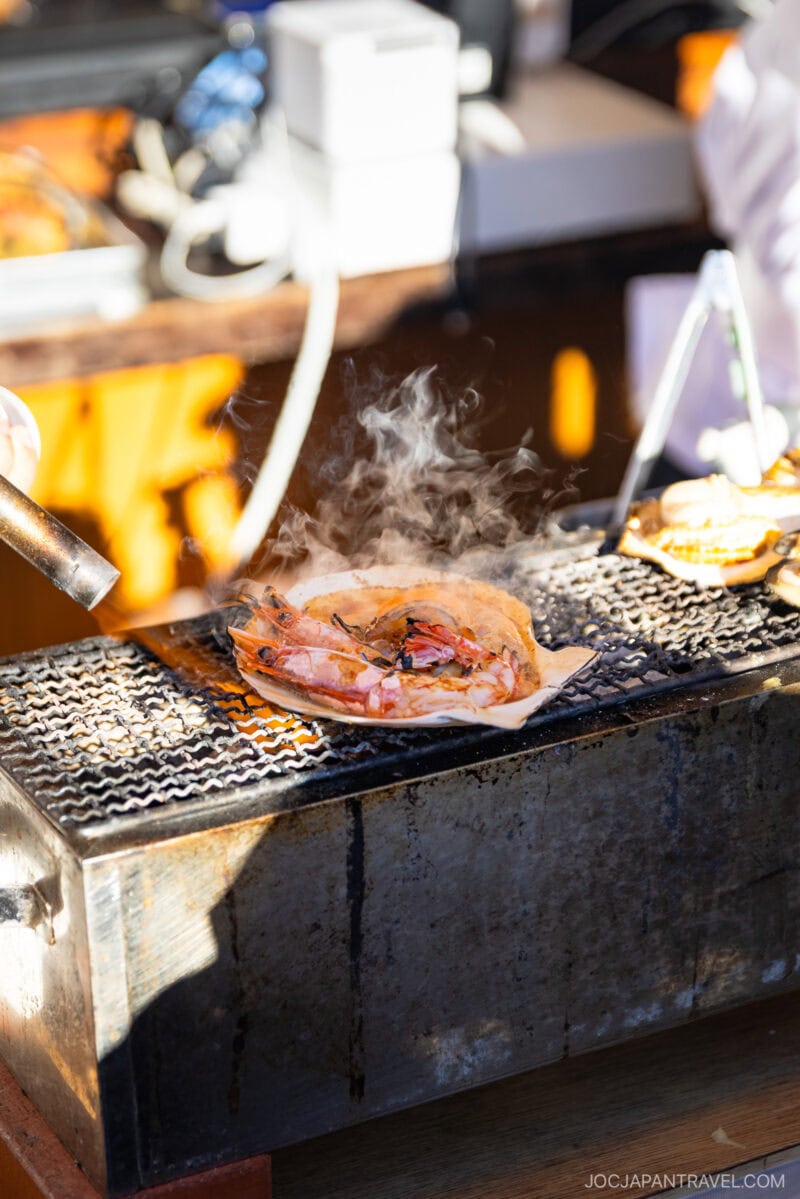 At the Tsukiji Outer Market in Tokyo, a seafood dish with shrimp and shellfish is grilled over an open flame on a metal grate, steam rising as cooking equipment fills the background at this lively outdoor food stall.