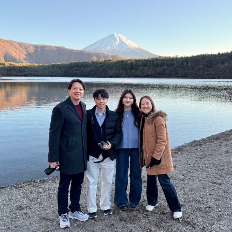 Four people stand together and smile by a calm lake with trees and Mount Fuji in the background under a clear sky. Two are holding cameras, and everyone is dressed warmly in coats.