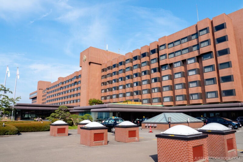 A large, modern brick hotel building with multiple floors and rows of windows stands against a blue sky—an ideal choice for those seeking where to stay in Japan. Cars are parked near the entrance, with small brick structures in the foreground.