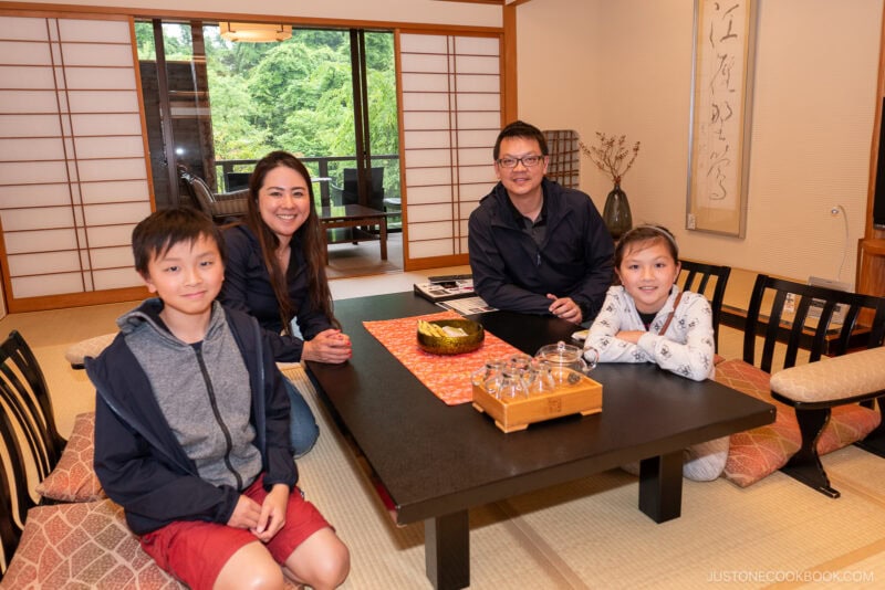 A family of four sits around a low table in a traditional Japanese-style room with tatami mats and shoji screens—an inviting glimpse for anyone wondering where to stay in Japan. They are smiling at the camera, sharing tea together.