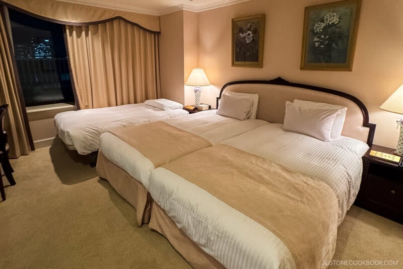Spacious hotel room—ideal if you're wondering where to stay in Japan—with two twin beds joined together, a single bed near the window, cozy beige and cream bedding, bedside tables with lamps, framed artwork, and stunning city lights outside.