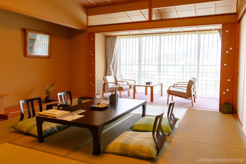 A traditional Japanese tatami room is a serene option for where to stay in Japan, featuring a low table, floor cushions, and chairs by a large window. A flower arrangement and books grace the table amid soft lighting and calm ambiance.