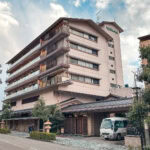 Where to Stay in Japan and What to Expect A multi-story building with balconies and traditional Japanese elements—perfect for those seeking where to stay in Japan. A van is parked in front, while trees and a sidewalk line the street under a cloudy sky.