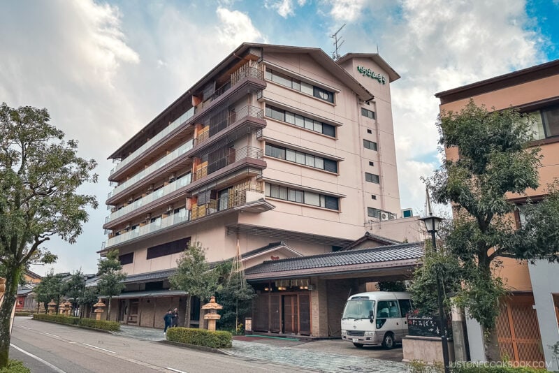 A multi-story building with balconies and traditional Japanese elements&mdash;perfect for those seeking where to stay in Japan. A van is parked in front, while trees and a sidewalk line the street under a cloudy sky.