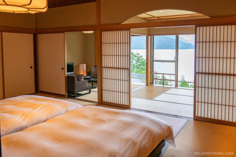 A serene Japanese-style room with tatami mats, two futon beds, sliding shoji doors, a sitting area, and large windows offering lake and mountain views—an ideal choice for those searching where to stay in Japan.
