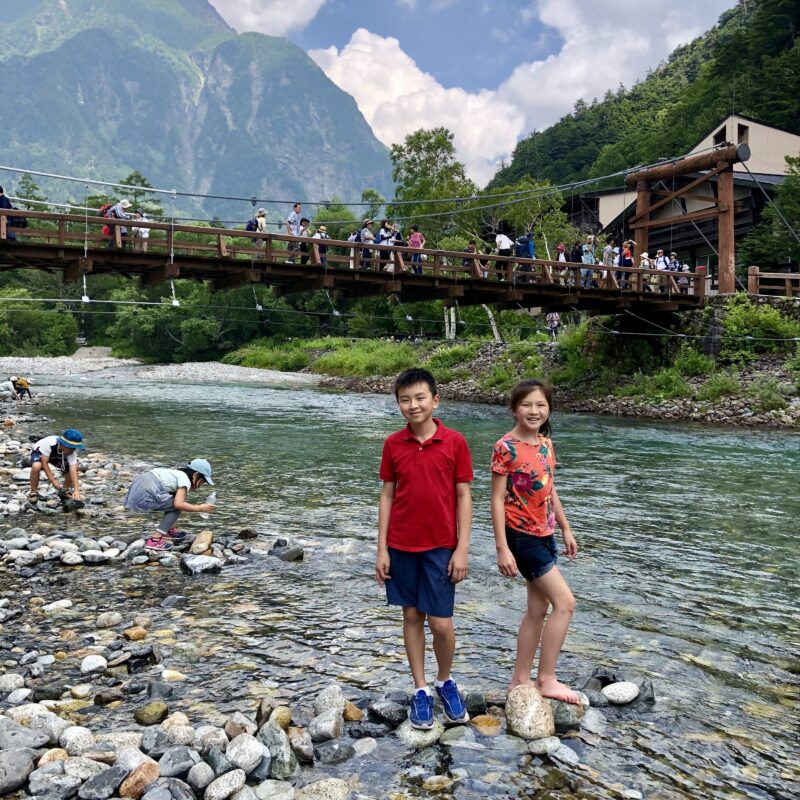 Two children stand on rocks in a clear river, smiling at the camera during their Japan travel. People cross a wooden bridge behind them, with green mountains and trees in the background under a partly cloudy sky.