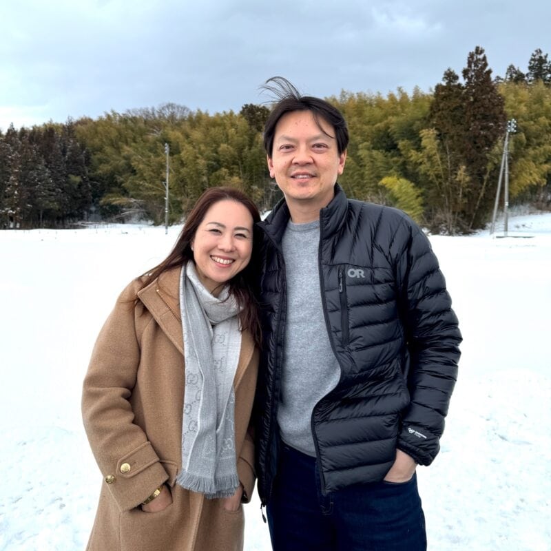 A smiling woman in a beige coat and scarf stands next to a smiling man in a black jacket. They are outdoors in a snowy landscape with trees and a cloudy sky in the background.