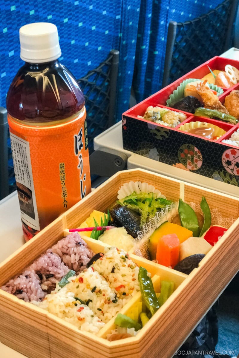 A bento box meal rests on a train tray table, featuring rice, vegetables, and pickles beside a bottle of tea—an experience often enjoyed by travelers with Japan train tickets. Another colorful bento and blue patterned seats appear in the background.