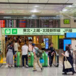 Japan Train Travel Guide: Tickets, IC Cards, and Shinkansen Basics People pass through ticket gates in a busy Japanese train station, scanning their Japan train tickets as illuminated signs above display schedules and destinations. The well-lit area bustles with travelers carrying bags.