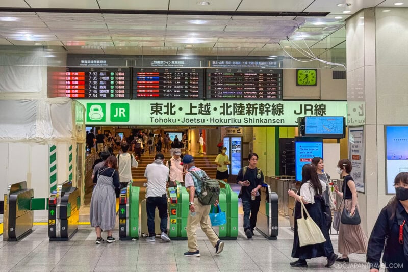 People pass through ticket gates in a busy Japanese train station, scanning their Japan train tickets as illuminated signs above display schedules and destinations. The well-lit area bustles with travelers carrying bags.