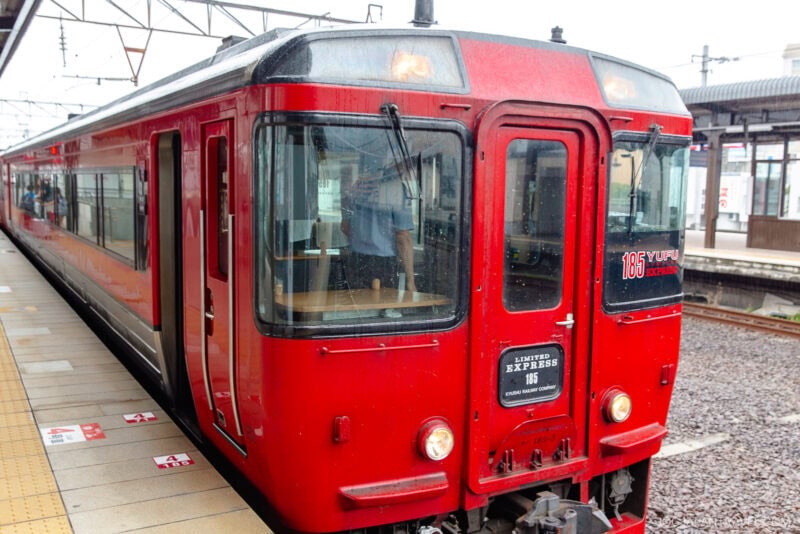 A red limited express train labeled Yufu 185 is stopped at a station platform on a cloudy day, with overhead wires and part of the station building visible in the background—an inviting scene for travelers with Japan train tickets.