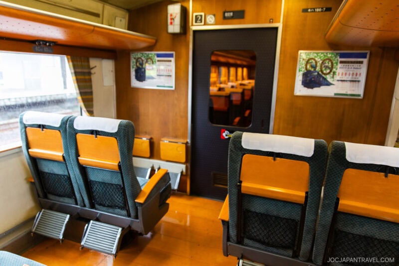 Interior of a train with green and orange seats, footrests, and a wooden-paneled wall. Posters highlight Japan train tickets, while a window and closed door to another car complete the cozy atmosphere.